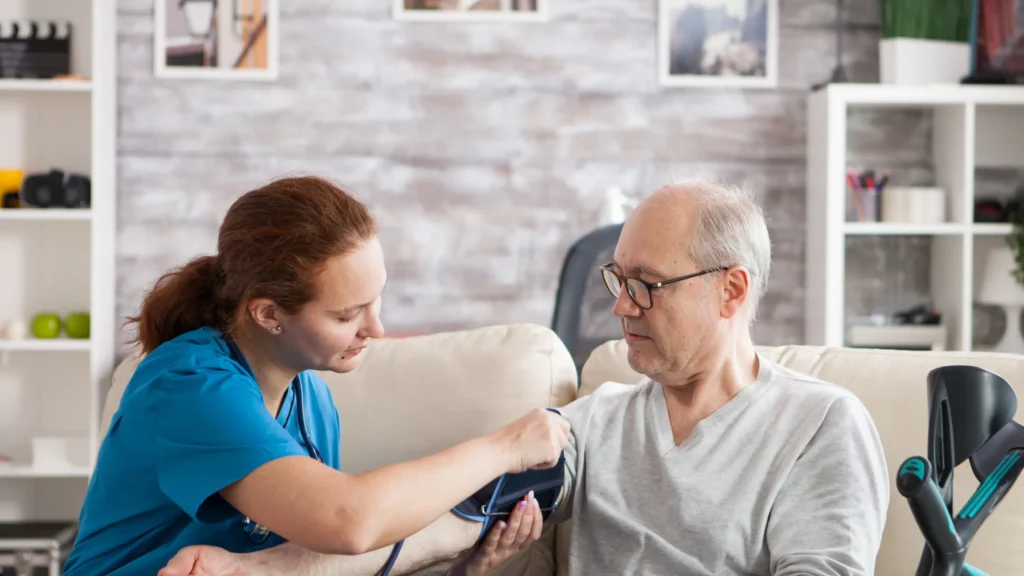 Caregiver checking a senior man's blood pressure on a living room couch during a professional home care visit